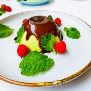 A green panna cotta made with pandan leaves covered in melted chocolate garnished with raspberry and mint leaves