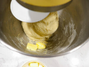 butter is added 1 piece at a time to a mixing bowl of a stand mixer to make fluffy dinner rolls