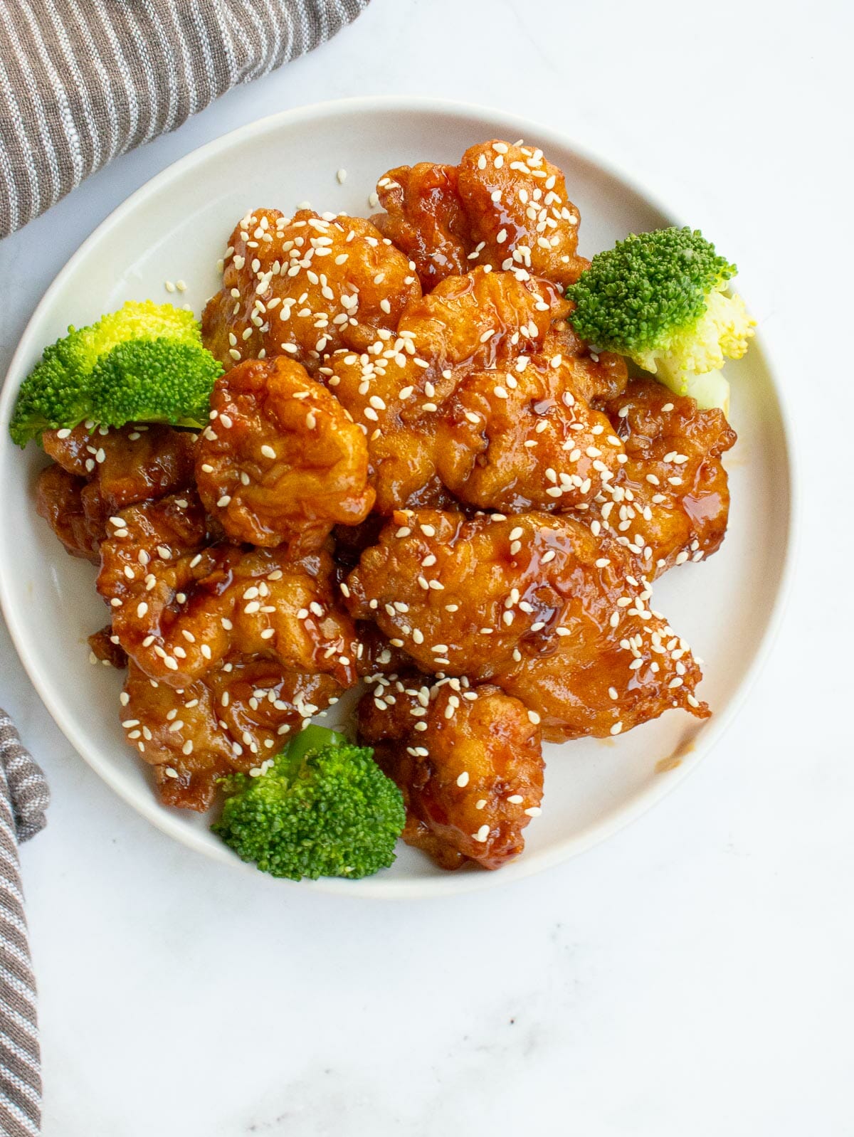 sesame chicken and steamed broccoli put on a white plate next to a brown stripe towel