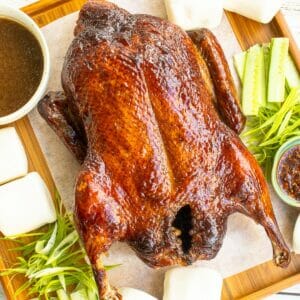 a whole roasted duck put on a wooden board next to slices of cucumber and green onion.