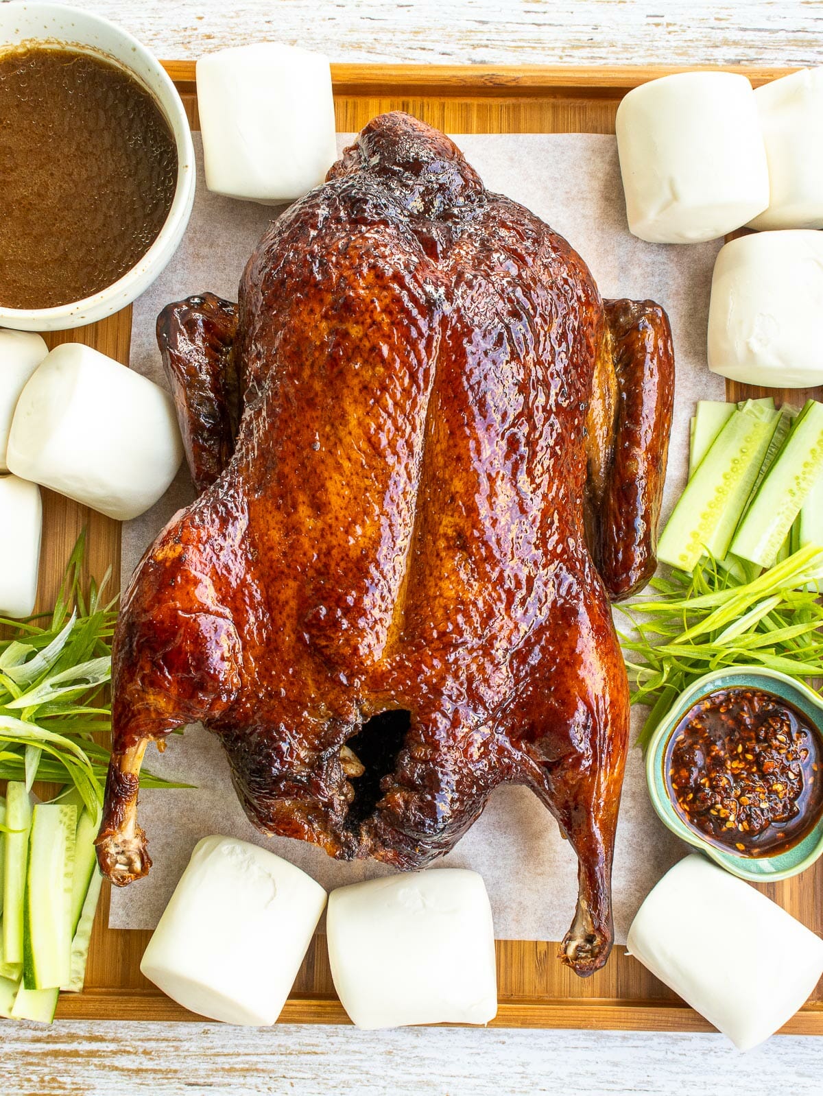 a whole Cantonese-style oven roasted duck put on wooden platter with bao, cucumber, green onion, sauce, and spicy sauce.