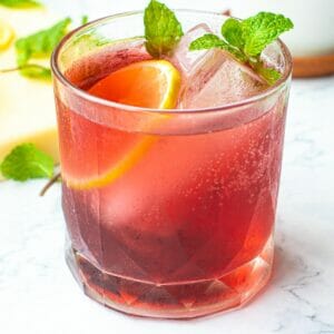 a cup of cranberry spritzer with 2 cubes of ice, slice of lemon and mint leaves