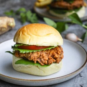 a burger that includes crispy chicken cutlets, assorted herbs, tomato slice put on a white plate