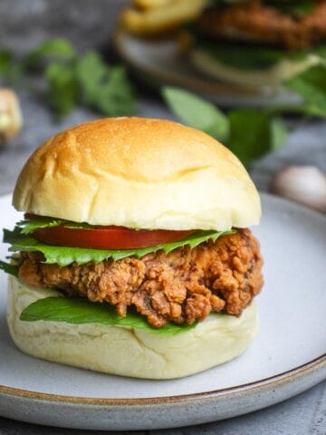 a burger that includes crispy chicken cutlets, assorted herbs, tomato slice put on a white plate